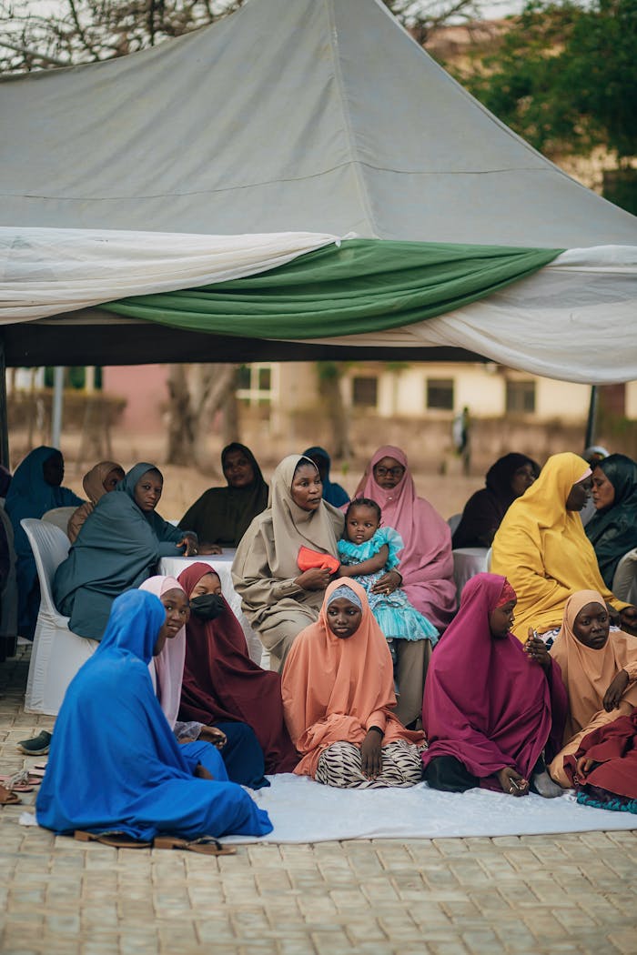gallery-2 Women and children in colorful attire sit outdoors under a canopy at a community event.